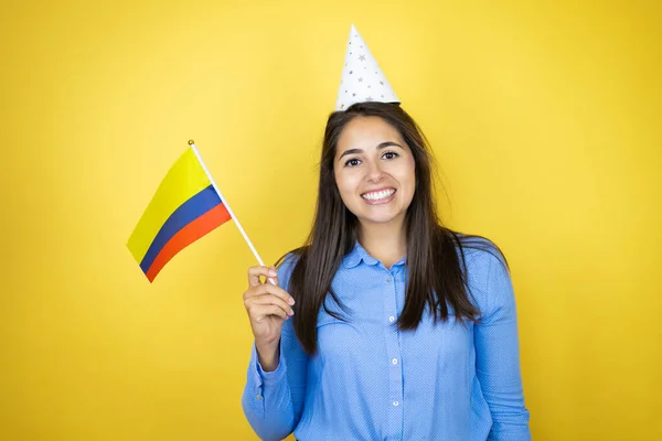 若いです白人女性の誕生日の帽子を身に着けています孤立した黄色の背景笑顔とコロンビアの旗を保持 — ストック写真