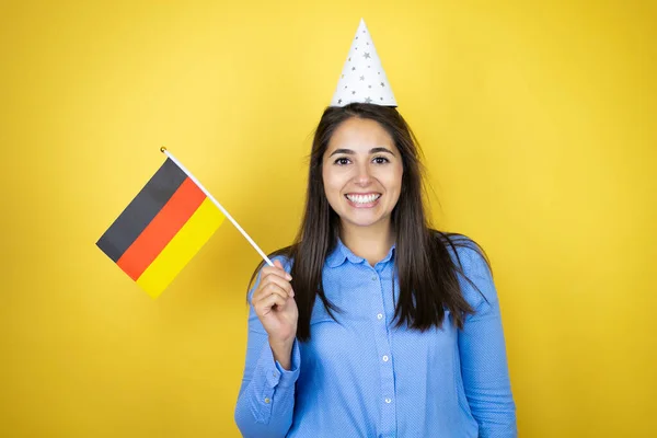 若いですCaucasian女性身に着けています誕生日帽子上の隔離された黄色の背景笑顔と保持ドイツのフラグ — ストック写真
