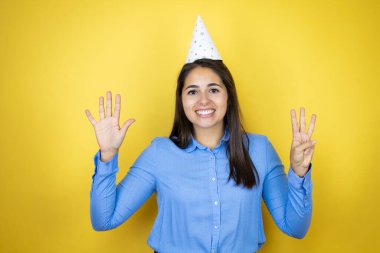 İzole edilmiş sarı arka planda doğum günü şapkası takan genç beyaz kadın sekiz numaralı parmaklarıyla gülümseyerek kendinden emin ve mutlu görünüyor.