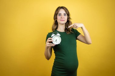 Güzel, esmer, hamile bir kadın izole edilmiş sarı arka plan üzerinde bebek bekliyor. Elinde bir saat tutarken ve doğrulturken şaşırmış.