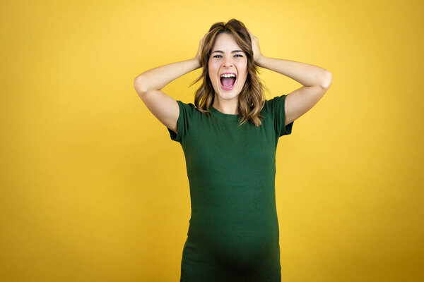 Молодая красивая брюнетка беременная ожидает ребенка на изолированном желтом фоне сумасшедший и напуганный руками на голове, боится и удивлен шока