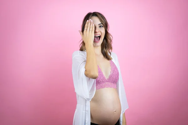 若いです美しいですブルネット女性予想赤ちゃん以上に孤立したピンクの背景カバー1目に手で 自信を持って笑顔上の顔と驚きの感情 — ストック写真