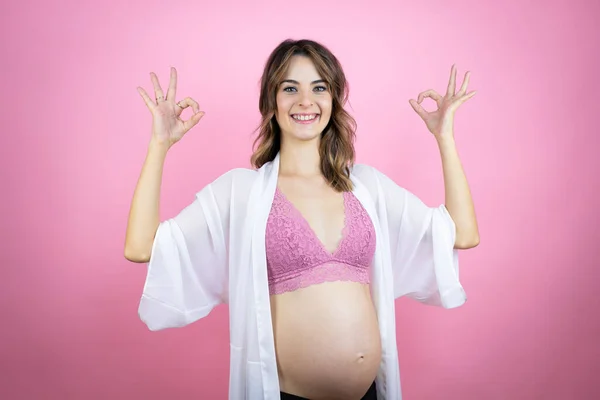 若いです美しいですブルネット女性妊娠予想赤ちゃん以上隔離されたピンクの背景をしますOk Signとともに指と笑顔 優れたシンボル — ストック写真