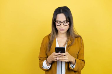 İzole edilmiş sarı arka plan üzerine blazer ceket giyen güzel bir kadın telefonuyla mesajlaşıyor.