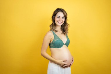 Güzel, esmer, hamile bir kadın, izole edilmiş sarı arka planda bebek bekliyor. Mutlu bir yüzü var, gülümsüyor ve kendine gülerek dişlerini gösteriyor.