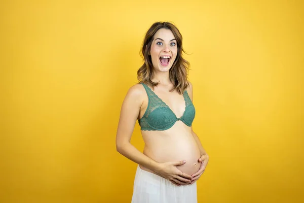 若いです美しいですブルネット女性妊娠期待しています赤ちゃん上の隔離された黄色の背景とともに幸せな顔立ちと驚いています自信を持って笑顔ショー歯 — ストック写真