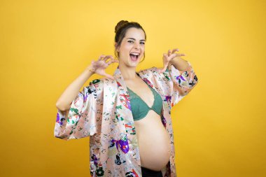Genç, güzel bir kadın hamile, izole edilmiş sarı arka planda pijama giyip bebek bekliyor. Komik bir şekilde pençe hareketi yapıyor, saldırgan ve seksi bir ifade.
