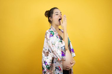 Genç, güzel bir kadın hamile, izole edilmiş sarı arka planda pijama giyiyor. Elle ağzını kapatmaktan sıkılmış. Huzursuz ve uykulu..