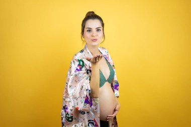 Güzel hamile bir kadın, izole edilmiş sarı arka planda pijama giymiş bebek bekliyor. Kameraya bakıyor ve havada öpücükler savuruyor. Güzel ve seksi. Aşk ifadesi.