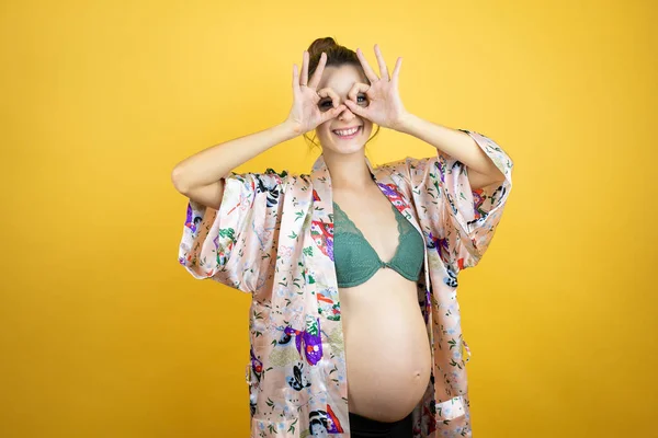 若いです美しいです女性妊娠期待赤ちゃん身に着けていますPajama上の隔離された黄色の背景行いますOkジェスチャーショックで笑顔 目を通して指 — ストック写真