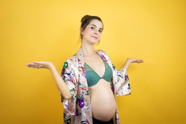 Genç, güzel bir kadın hamile, izole edilmiş sarı arka planda pijama giyiyor ve elleri ve kolları havada, kafası karışmış bir ifade takınıyor.