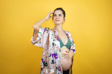 Genç, güzel bir kadın hamile, izole edilmiş sarı arka planda bebek pijaması giyiyor. Soruyu merak ediyor. Şüpheyle karışık, kafa kafaya düşünerek.