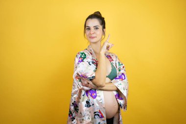 Genç, güzel bir kadın hamile, izole edilmiş sarı arka planda pijama giyiyor ve elleri ve parmakları kenarı işaret ediyor.