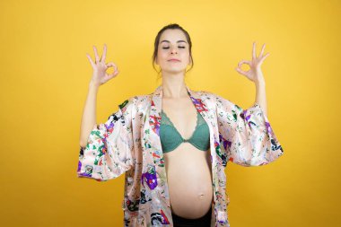 Genç, güzel bir kadın hamile, izole edilmiş sarı arka planda pijama giyiyor. Gözleri kapalı gülümsüyor. Parmaklarıyla meditasyon hareketi yapıyor.