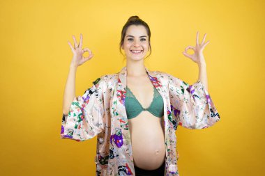 Genç, güzel bir kadın hamile, izole edilmiş sarı arka planda pijama giyiyor. Tamam işareti yapıyor, gülümsüyor, mükemmel bir sembol.