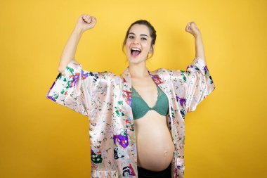 Genç, güzel bir kadın hamile, izole edilmiş sarı arka planda pijama giyiyor. Çok mutlu ve heyecanlı. Kollarını kaldırmış, gülümseyen ve başarı için bağıran bir jest yapıyor..
