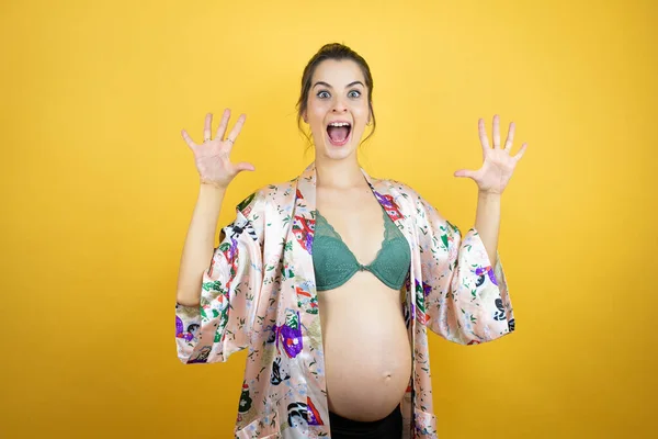 若いです美しいです女性妊娠期待赤ちゃん身に着けていますPajama上の隔離された黄色の背景ショーと指差しますと指番号10とともに笑顔自信と幸せ — ストック写真