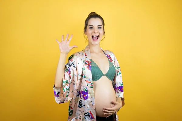 若いです美しいです女性妊娠期待赤ちゃん身に着けていますPajama上の隔離された黄色の背景ショーと指差しますと指番号5とともに笑顔自信と幸せ — ストック写真