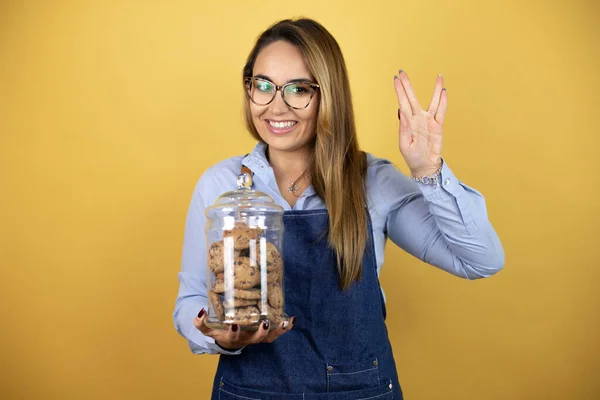 Young Hispanic Woman Wearing Baker Uniform Holding Cookies Jar Doing — Stock Photo, Image