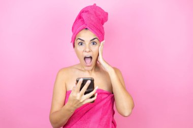 Banyodan sonra duş havlusu giyen genç ve güzel bir kadın izole edilmiş pembe arka planda dikilip telefonda endişeli bir ifadeyle sohbet ediyor.
