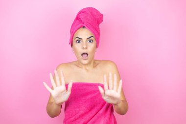 İzole edilmiş pembe arka planda duran duş havlusu giyen genç ve güzel bir kadın korku ifadesinden korkmuş ve dehşete düşmüş halde duruyor. El hareketlerini durdur, şok içinde bağır. Panik konsepti.