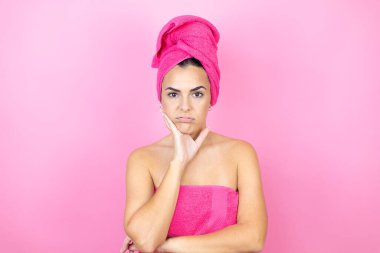 Banyodan sonra duş havlusu giyen genç ve güzel bir kadın izole edilmiş pembe arka planda durup kollarını kavuşturmaktan sıkılmış ve yorgun göründüğünü düşünüyor.