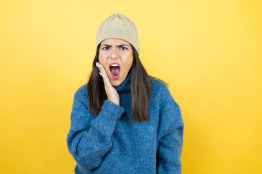 Mavi süveter ve yün şapka giyen genç ve güzel bir kadın diş ağrısı ya da diş hastalığından dolayı ağızlarına acı dolu bir ifadeyle dokunuyor.