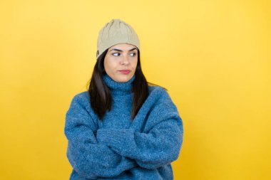 Mavi süveter ve yün şapka giyen genç ve güzel bir kadın kollarını kavuşturmaktan sıkılmış ve yorgun görünüyor.