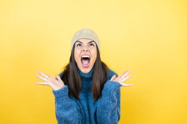 Mavi süveter ve yün şapka giyen, kolları ve elleri havada, kafası karışmış genç ve güzel bir kadın.