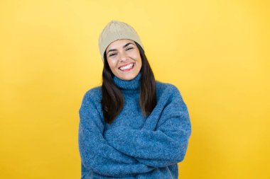 Mavi süveter ve gülen yüzlü yün şapka giyen genç ve güzel bir kadın kollarını kavuşturmuş gülümseyerek ayakta duruyor.