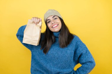 Mavi süveter giyen, yün şapka takan, gülümseyen, kağıt torba tutan güzel bir kadın.