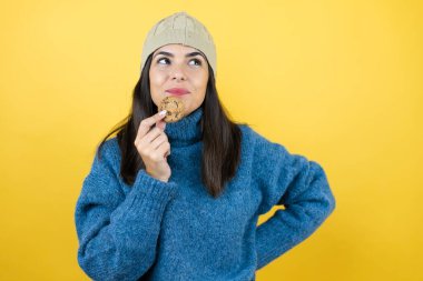 Mavi süveter ve yün şapka giyen, mutlu yüzüyle çikolatalı kurabiye yiyen, kendine güvenen, gülümseyen, dişlerini gösteren genç ve güzel bir kadın.