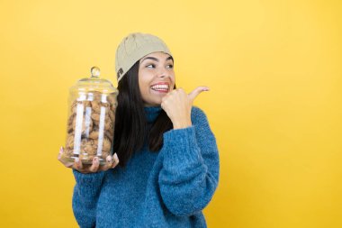 Mavi süveter ve yün şapka giyen, elinde çikolata parçacıklı kurabiye kavanozu tutan, gülümseyen ve baş parmağıyla yüzünü gösteren genç ve güzel bir kadın.