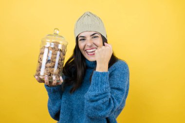 Mavi süveter ve yün şapka giyen, elinde çikolatalı kurabiye kavanozu tutan genç ve güzel bir kadın. Çok mutlu ve heyecanlı. Kollarını kaldırıp, gülümseyerek ve başarı için çığlık atarak kazanan bir jest yapıyor..