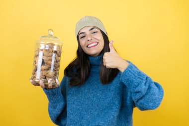 Mavi süveter ve yün şapka giyen, elinde çikolata parçacıklı kurabiye kavanozu tutan, gülümseyen ve baş parmağıyla işaret veren genç ve güzel bir kadın.