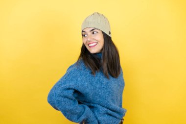 Mavi süveter ve yün şapka giyen genç ve güzel bir kadın kollarını kavuşturmuş ve kendinden emin bir şekilde yan tarafa bakıyor.