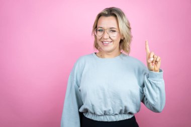 Pembe arka planda eşofman üstü giyen genç beyaz kadın bir numaralı parmaklarını gösterirken kendine güvenen ve mutlu bir şekilde gülümsüyor.