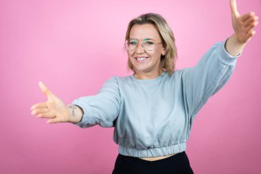 Pembe arka planda eşofman üstü giyen beyaz bir kadın kameraya sarılıp gülümsüyor.