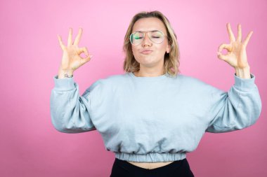 Pembe arka planda eşofman üstü giyen genç beyaz kadın rahatla ve gözleri kapalı gülümse parmaklarıyla meditasyon hareketi yapıyor.