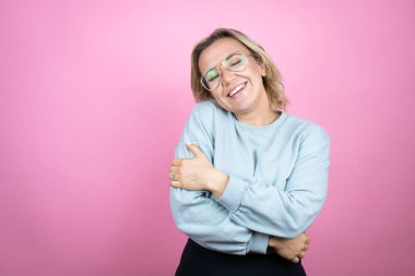 Pembe arka planda eşofman üstü giyen beyaz bir kadın. Kendine sarılıyor, mutlu ve pozitif, kendinden emin gülümsüyor.