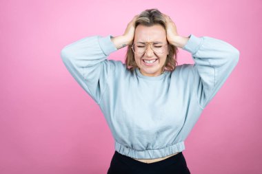 Pembe arka plan üzerine kazak giyen genç beyaz kadın baş ağrısı ve stresten muzdarip çünkü elleri başının üstünde acı ve migren var.