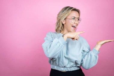 Pembe arka planda eşofman üstü giyen beyaz bir kadın elinde olan bir şeyi gösterip işaret ediyor.