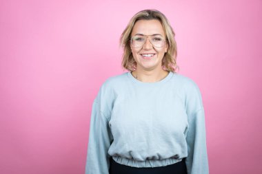 Pembe arka planda eşofman üstü giyinmiş, gülümseyen, kendine güvenen bir gülümsemeyle gülümseyen beyaz bir kadın.