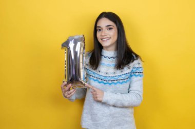 Sarı arka planda günlük süveter giyen beyaz bir kadın bir numaralı balonu tutuyor. Kutlama konsepti