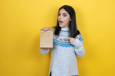 Sarı arka planda günlük süveter giyen genç beyaz kadın. Gülümsüyor ve elinde kağıt torba ile onu işaret ediyor.