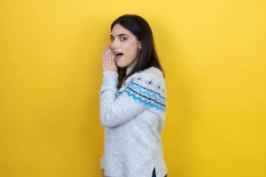 Sarı arka plan üzerine günlük süveter giyen genç beyaz bir kadın. Ağız üstüne el, gizli dedikodular, kötü niyetli konuşmalar.