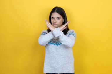 Sarı arka plan üzerine günlük süveter giyen beyaz bir kadın. Reddedilmiş yüz ifadesi. Kollarını kavuşturup negatif işaret veriyor.