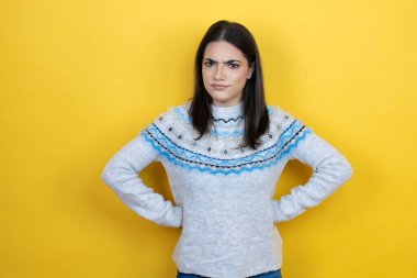 Sarı arka plan üzerine sıradan süveter giyen genç beyaz kadın kuşkucu ve gergin, yüzünde tasvip etmeyen bir ifade var.