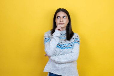 Sarı arka plan üzerine günlük süveter giyen beyaz bir kadın ciddi bir yüz ifadesi olan eli çenesinde soru soran, kafa karıştırıcı bir fikir hakkında düşünceli bir kadın.