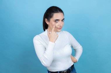 Mavi arka planda beyaz süveter giyen genç beyaz bir kadın. Gözünü dikip jestini izliyor. Şüpheli bir ifade.
